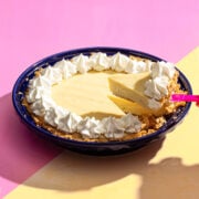A slice of creamy yellow pie with a graham cracker crust and whipped cream topping is being lifted from a dark blue pie dish, set on a pink and yellow background.