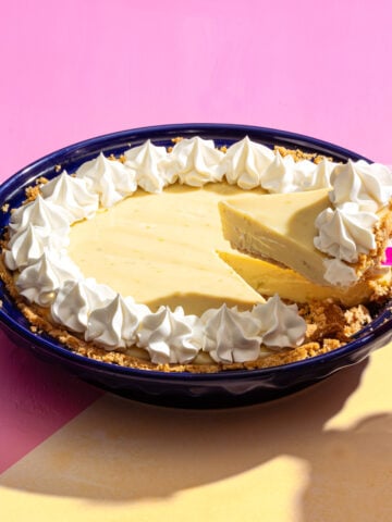 A slice of creamy yellow pie with a graham cracker crust and whipped cream topping is being lifted from a dark blue pie dish, set on a pink and yellow background.