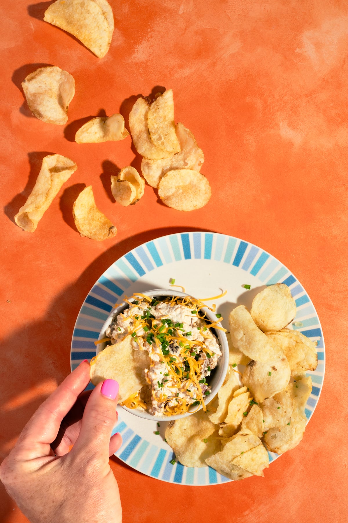 A hand with pink nail polish dips a potato chip into a bowl of creamy dip, surrounded by more chips on a striped plate and scattered on an orange surface.