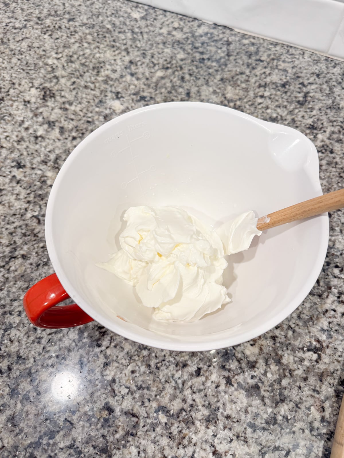 A white mixing bowl with a red handle contains a dollop of whipped cream and has a wooden spoon resting inside. The bowl sits on a granite countertop.