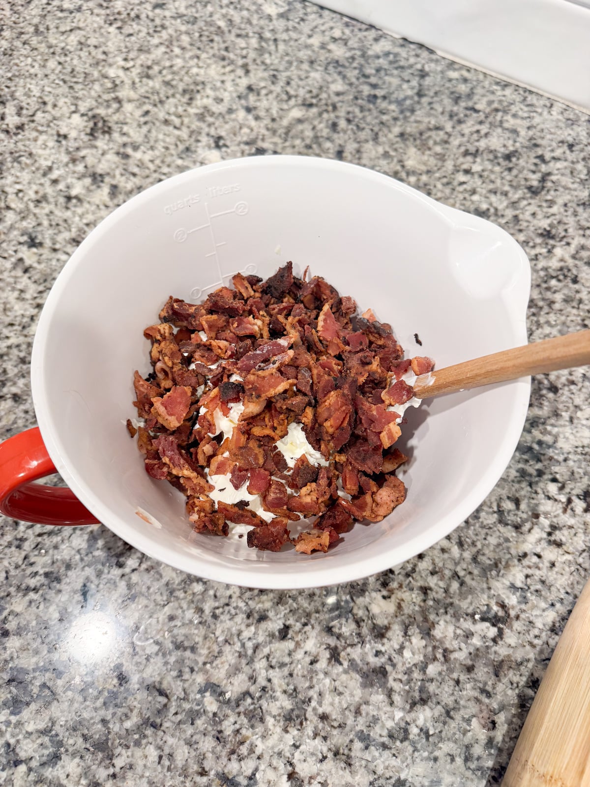 A white mixing bowl with a red handle contains chopped cooked bacon and cream cheese being mixed with a wooden spoon. The bowl is on a speckled granite countertop.