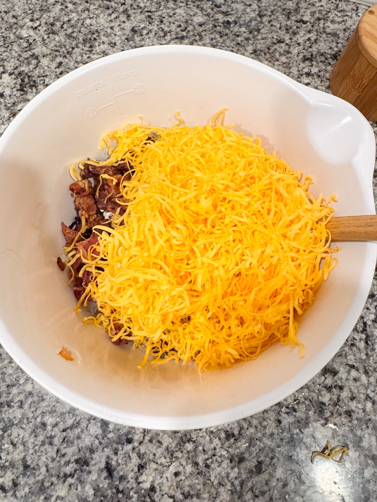 A white mixing bowl on a granite countertop contains shredded cheddar cheese and chopped cooked bacon, with a wooden spoon resting inside the bowl.