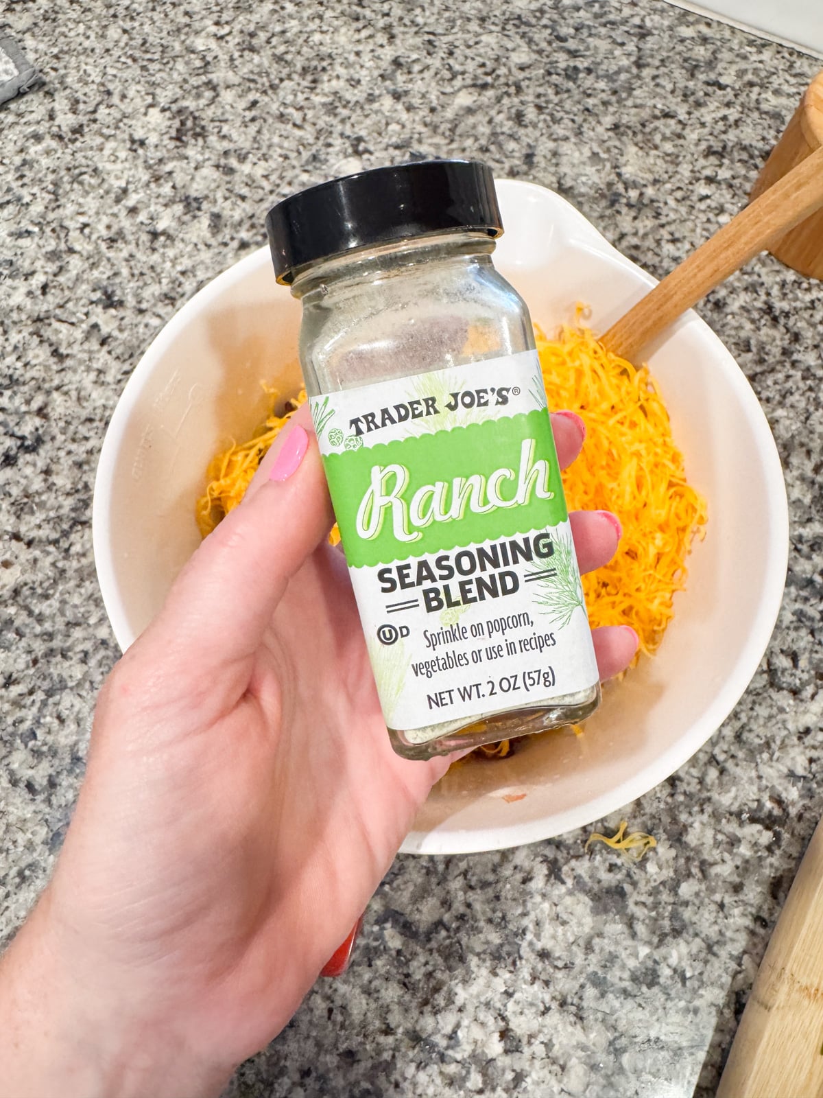 A hand holds a jar of Trader Joes Ranch Seasoning Blend over a white bowl filled with shredded orange cheese on a granite countertop.