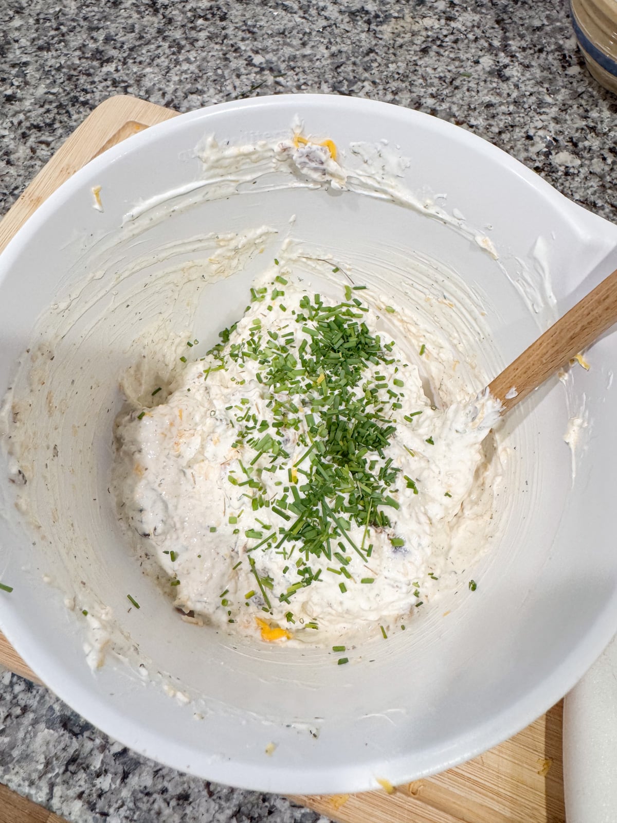 A white mixing bowl on a counter holds a thick, creamy batter with chopped chives on top and a wooden spoon resting in the mixture.