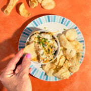 A hand dips a potato chip into a bowl of cheesy dip, garnished with chives, on a plate with more chips. The plate sits on an orange surface with extra chips scattered around.