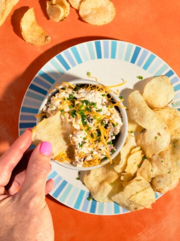 A hand dips a potato chip into a bowl of cheesy dip, garnished with chives, on a plate with more chips. The plate sits on an orange surface with extra chips scattered around.