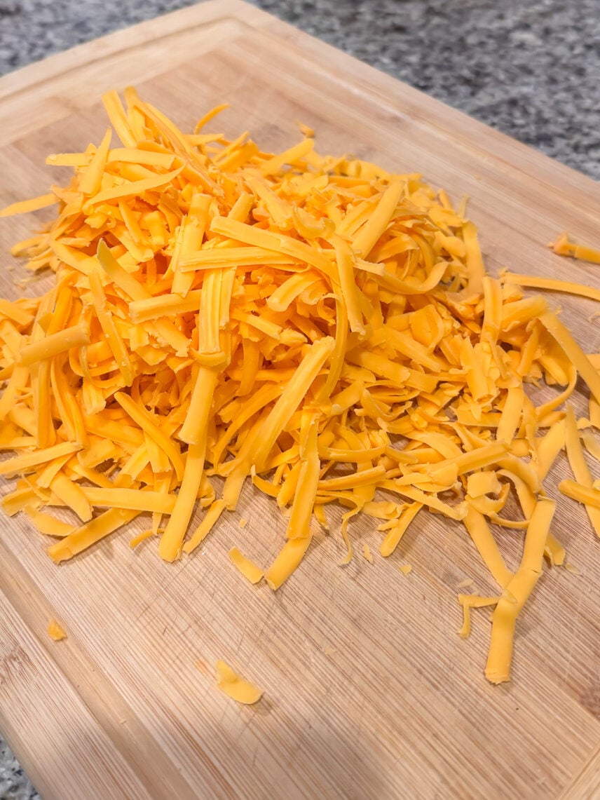 A pile of shredded cheddar cheese sits on a wooden cutting board, ready to add savory flavor to your next dish—perhaps a Croissant Breakfast Casserole with Sausage and Pimentos. In the background, a glass dish of cubed bread rests on a gray countertop, perfect for creating this comforting breakfast favorite.