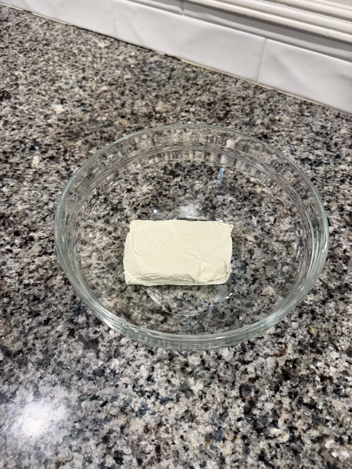 A glass bowl with a block of cream cheese sits on a granite countertop. White tiles are visible in the background.