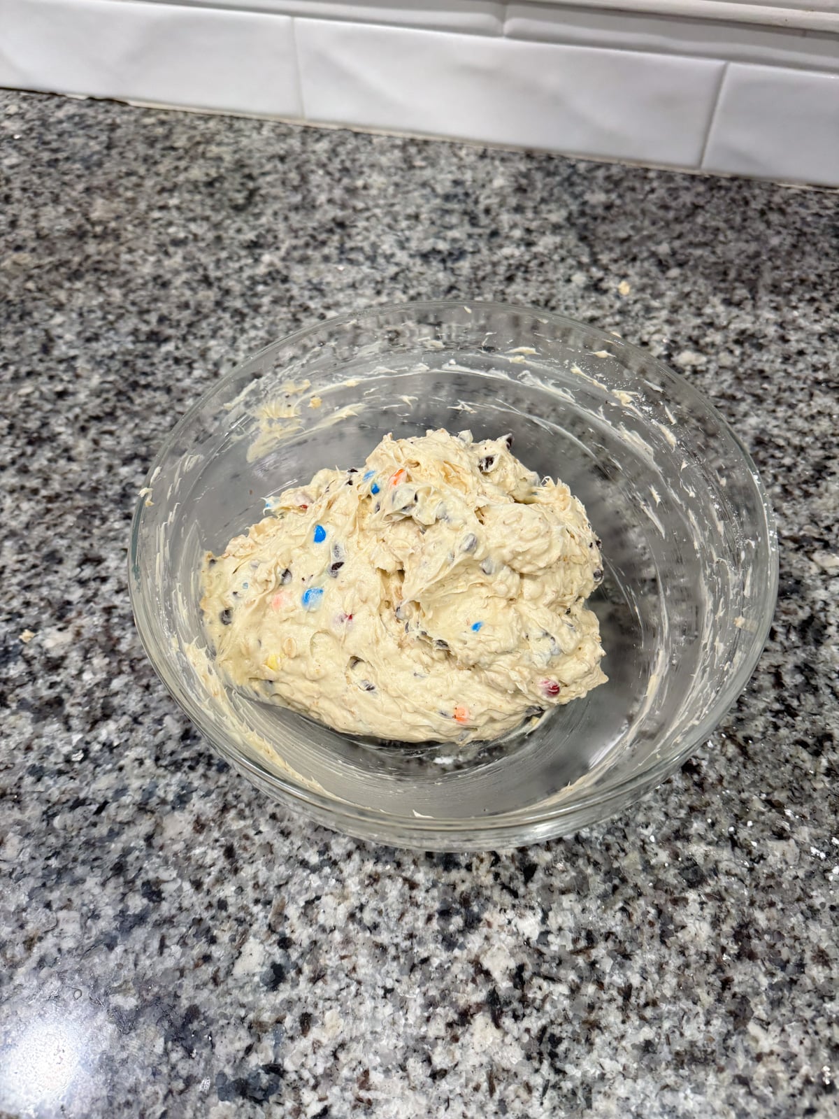 A glass bowl filled with cookie dough containing colorful candy pieces sits on a gray granite countertop with white tile backsplash in the background.