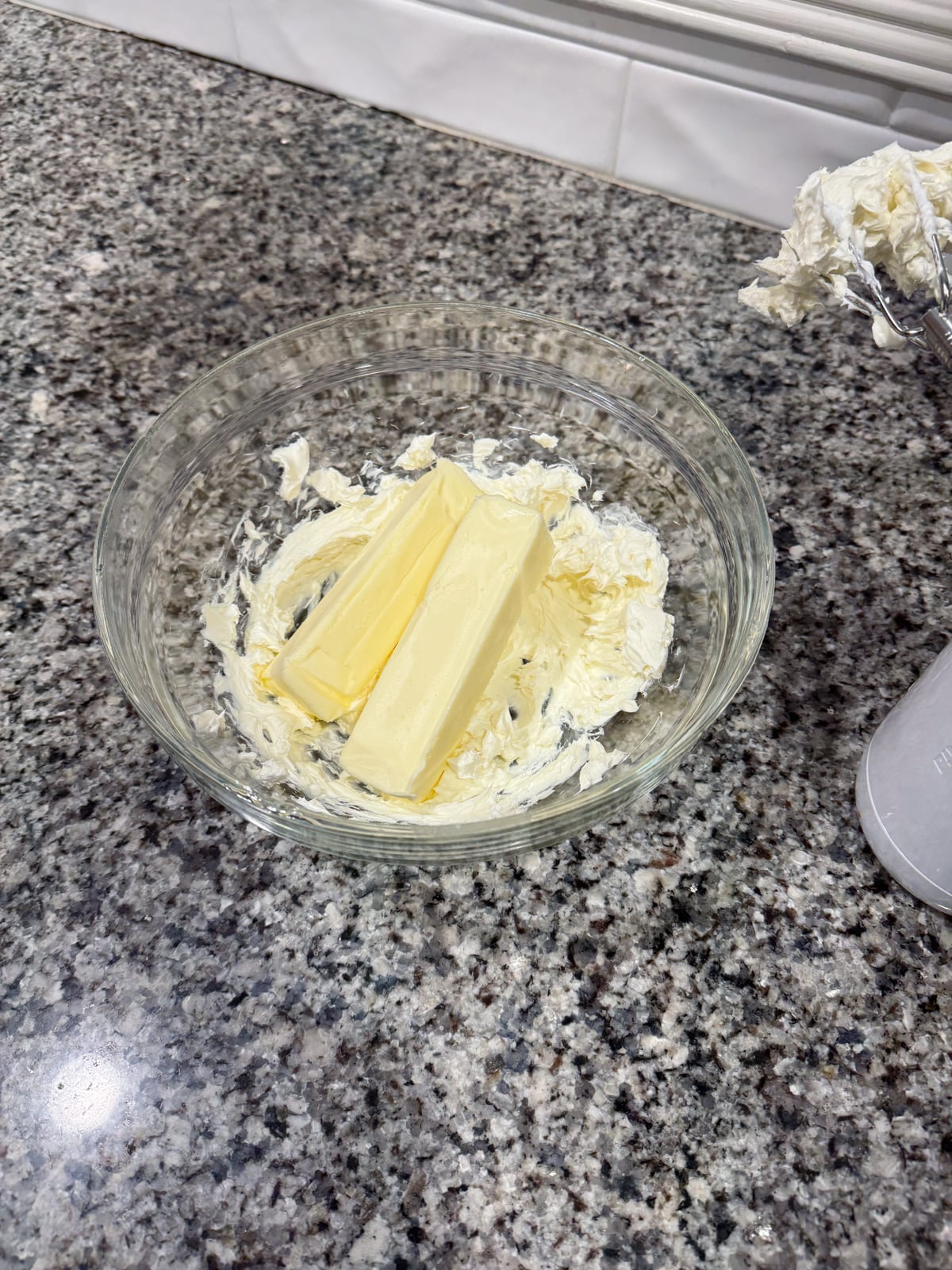 A glass bowl on a granite counter contains two sticks of butter and some creamed butter, with a hand mixer and beaters nearby.