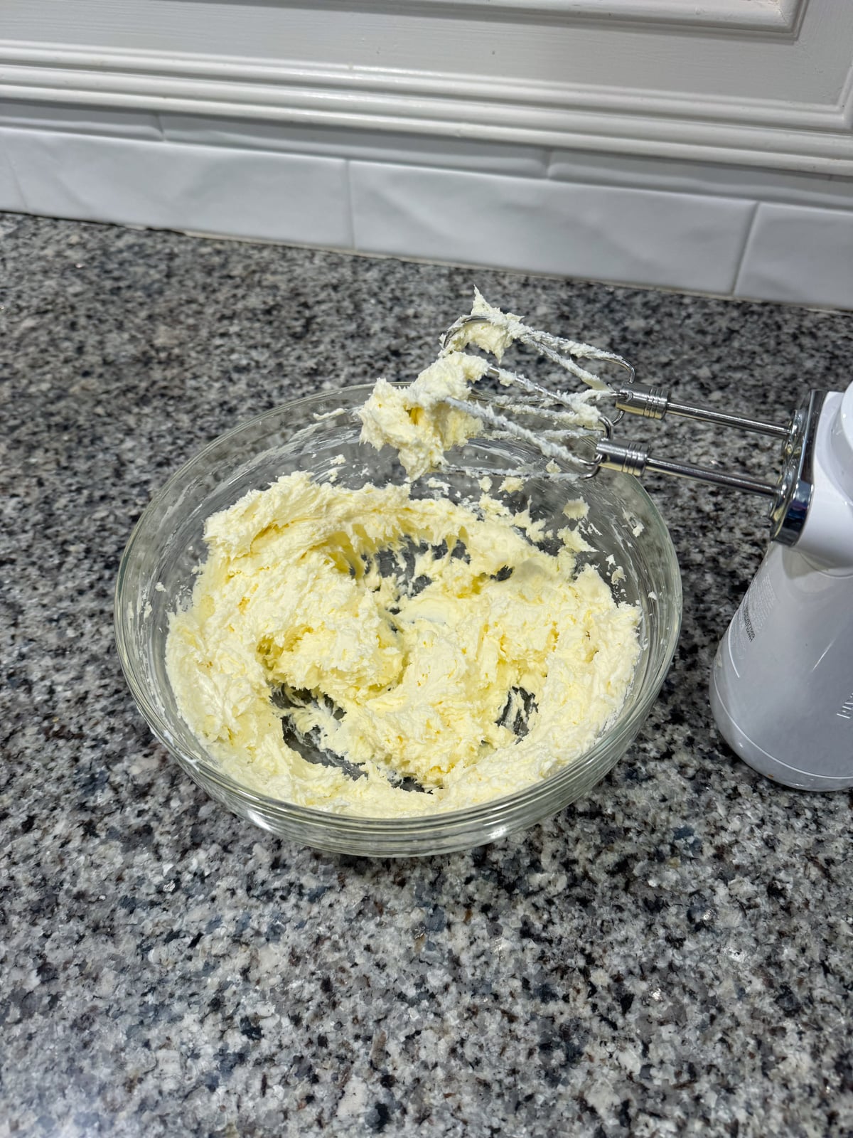 A glass bowl of creamed butter and sugar sits on a granite countertop beside a handheld electric mixer with some batter on its beaters. White tile backsplash is in the background.