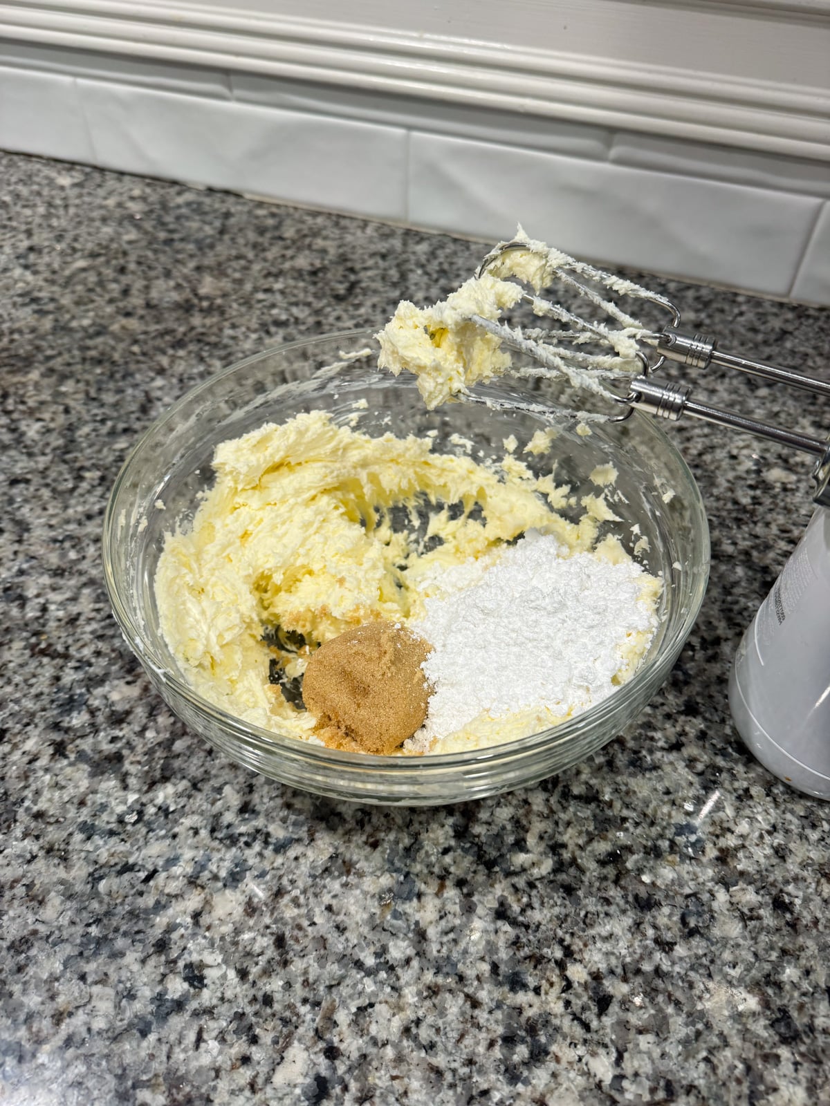 A glass bowl on a granite countertop contains creamed butter with brown and powdered sugar. An electric hand mixer rests on the bowls edge.