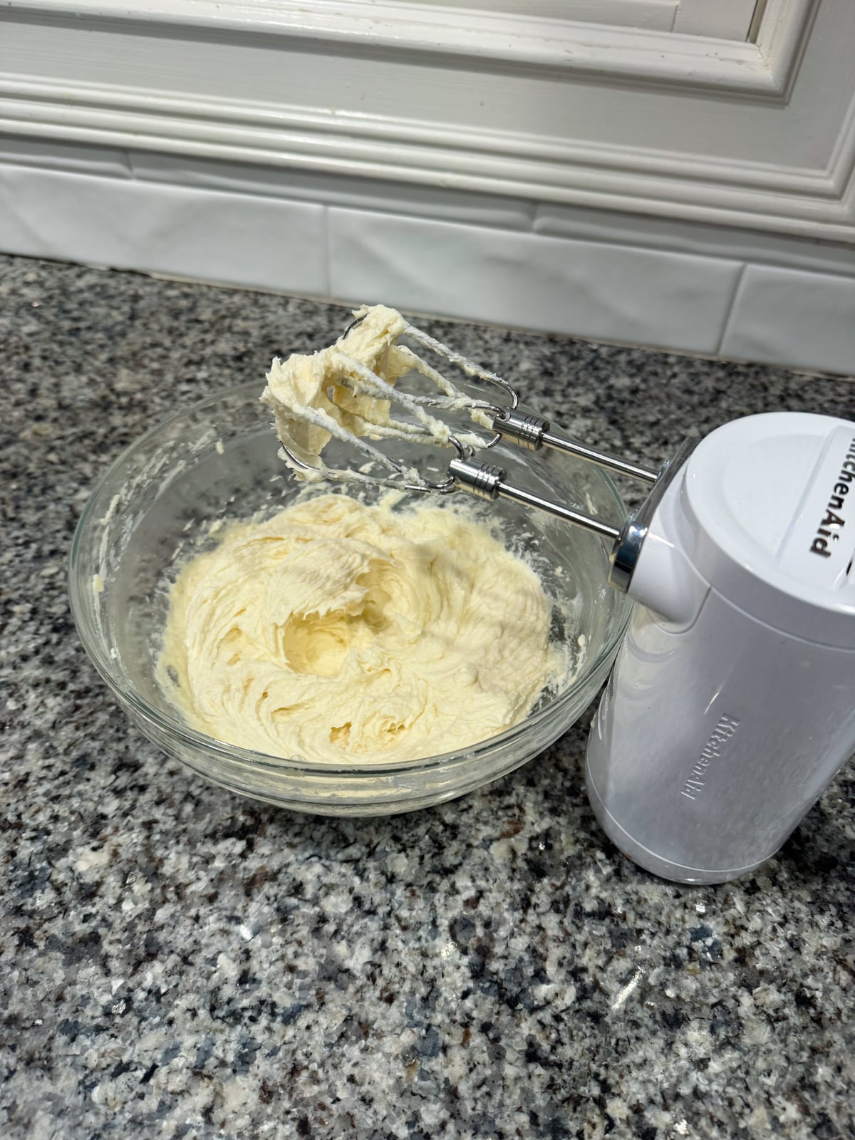 A glass bowl with creamy batter sits on a granite countertop. An electric hand mixer with batter on its beaters rests nearby. White tile backsplash and window frame are visible in the background.