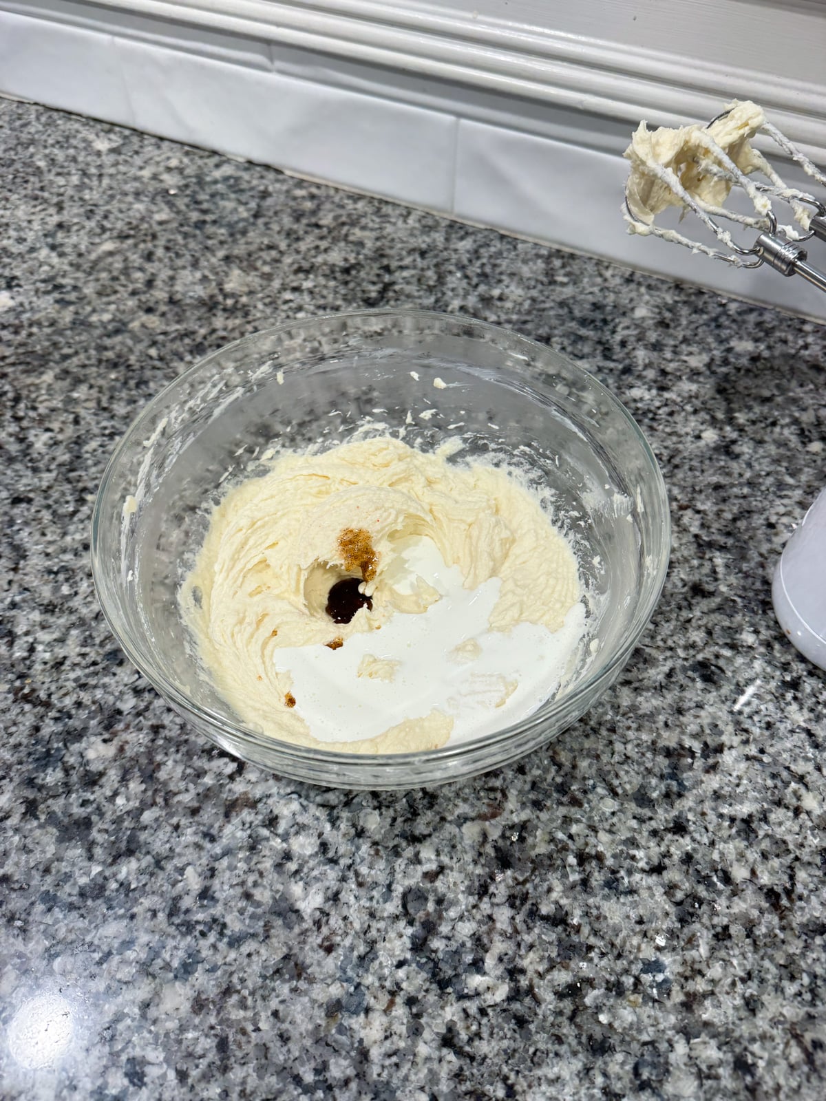 A glass bowl of partially mixed batter with vanilla extract and cream sits on a granite countertop. An electric mixer with batter on the beaters rests nearby.