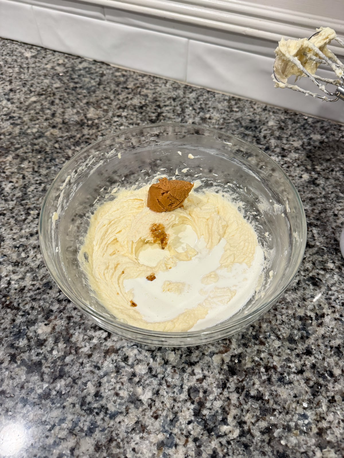 A glass bowl with creamy batter, a splash of milk, and a dollop of brown paste, likely vanilla or cinnamon, sits on a granite countertop. A hand mixer with batter on its beaters rests nearby.