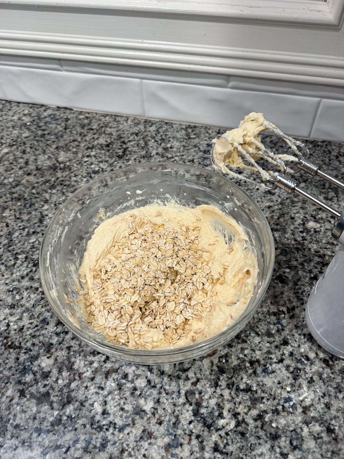 A glass mixing bowl with cookie dough and oats on top sits on a granite countertop. An electric hand mixer with dough-covered beaters rests beside the bowl. White tile backsplash is visible in the background.