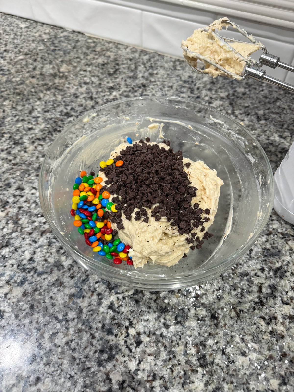 A glass bowl with cookie dough, mini chocolate chips, and colorful candy-coated chocolates on a granite countertop. An electric mixer with dough on the beaters rests nearby.