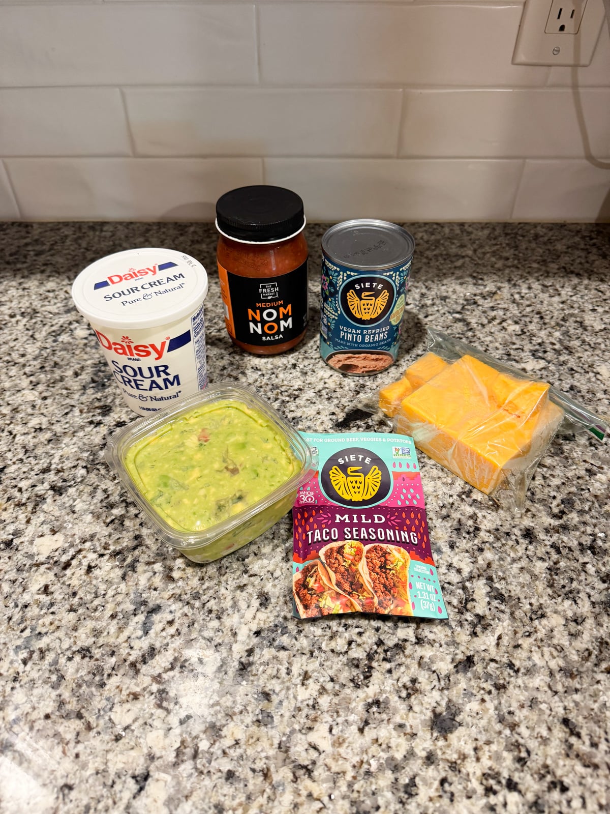Taco ingredients on a granite counter: sour cream, salsa, canned pinto beans, shredded cheddar cheese in a bag, guacamole in a container, and mild taco seasoning. White tile backsplash and outlet in the background.
