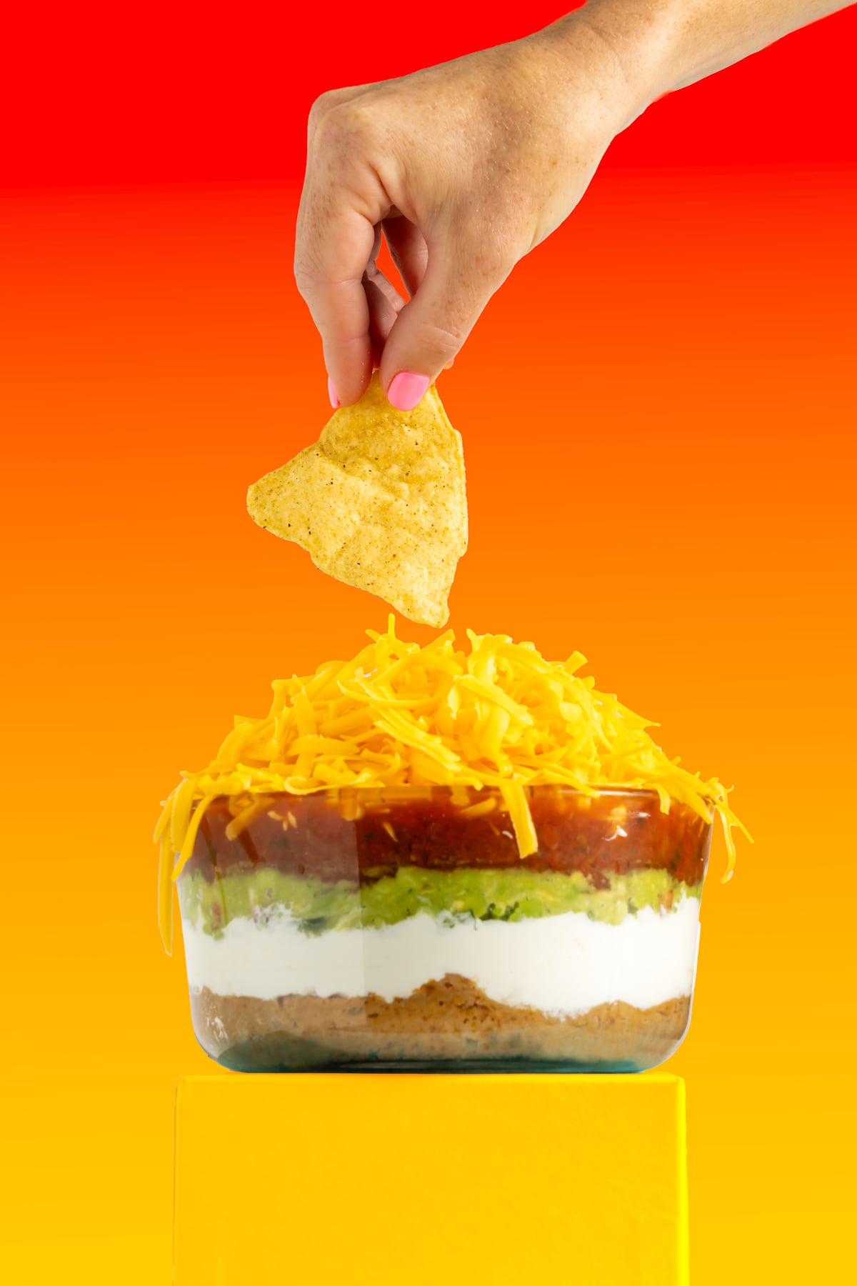 A hand holding a tortilla chip above a layered dip with cheese, salsa, guacamole, and sour cream in a glass bowl, against a vibrant orange and red background.
