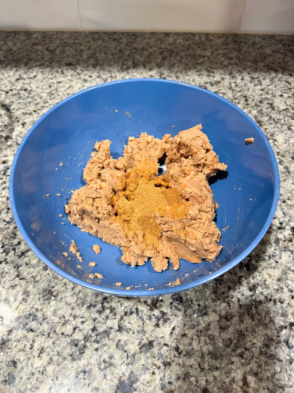 A blue bowl on a granite countertop contains mashed refried beans with a pile of taco seasoning on top, ready to be mixed.