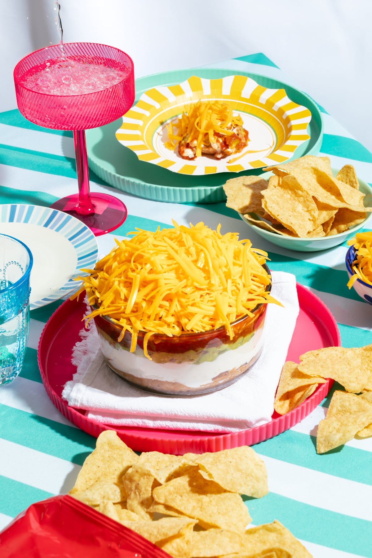 A table set with a layered dip topped with shredded cheese, tortilla chips, and a pink drink in a stemmed glass. Bright plates and a striped tablecloth add color to the scene.