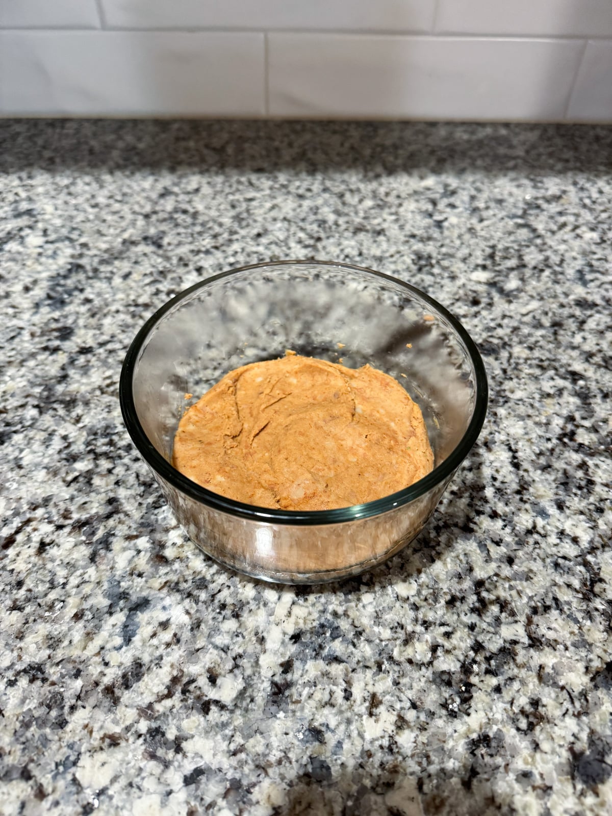 A small glass bowl filled with a light brown, creamy mixture sits on a speckled granite countertop in front of a white tiled backsplash.