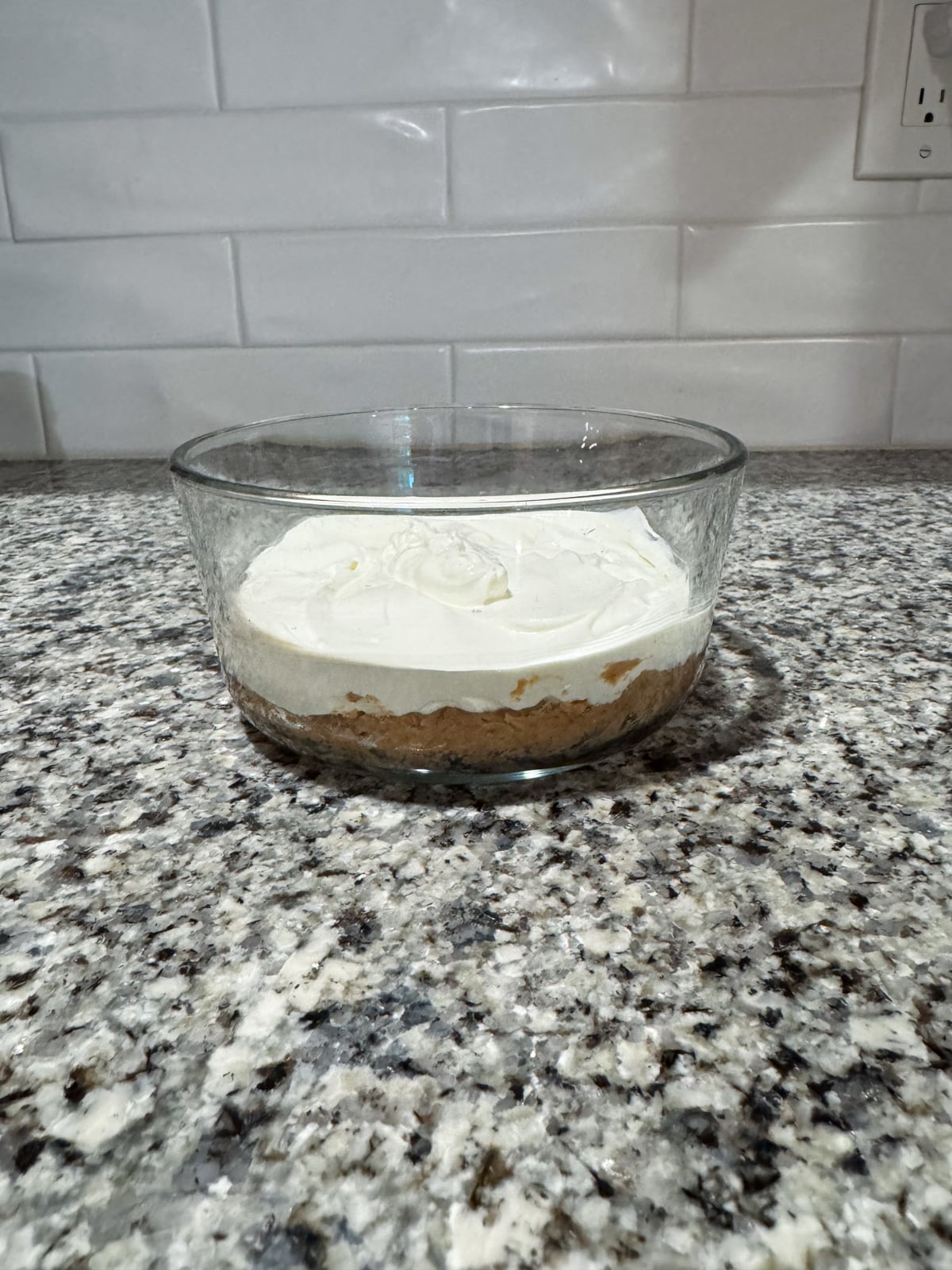 A glass bowl with a dessert featuring a crumbly brown base and a thick layer of creamy white topping sits on a speckled granite countertop with a white tile backsplash.
