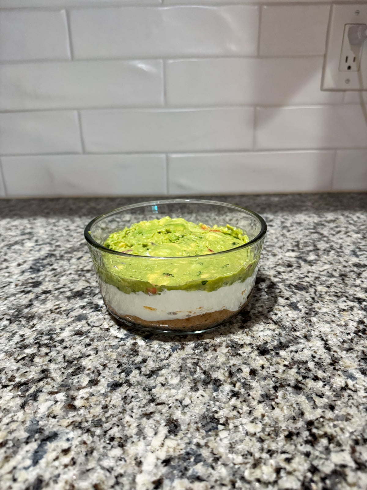 A glass bowl filled with layered dip, featuring a bottom brown layer, a middle white layer, and a top layer of chunky green guacamole, sits on a granite countertop in front of a white tiled backsplash.