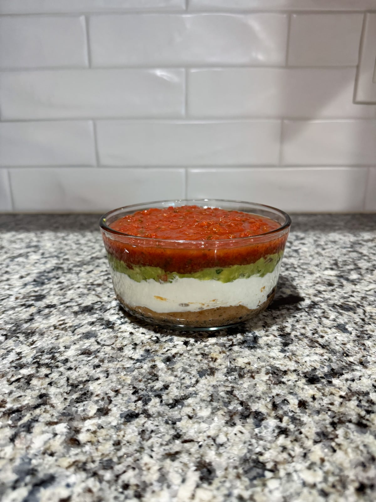 A glass bowl with layered dip containing salsa, guacamole, sour cream, and a brown base sits on a granite countertop in front of a white tiled backsplash.