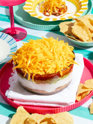 A bowl of seven-layer dip topped with shredded cheddar cheese sits on a pink tray, surrounded by tortilla chips and plates on a striped tablecloth.