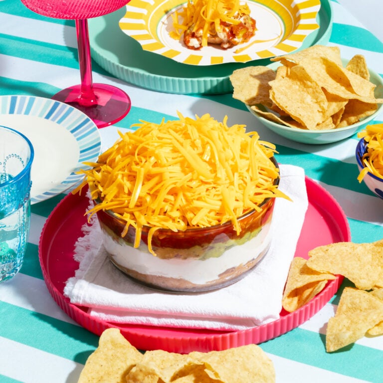 A bowl of seven-layer dip topped with shredded cheddar cheese sits on a pink tray, surrounded by tortilla chips and plates on a striped tablecloth.